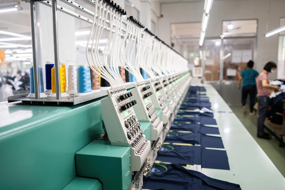 Máquina industrial de bordado em linha de produção da manufatura têxtil com fios coloridos e operação automatizada.